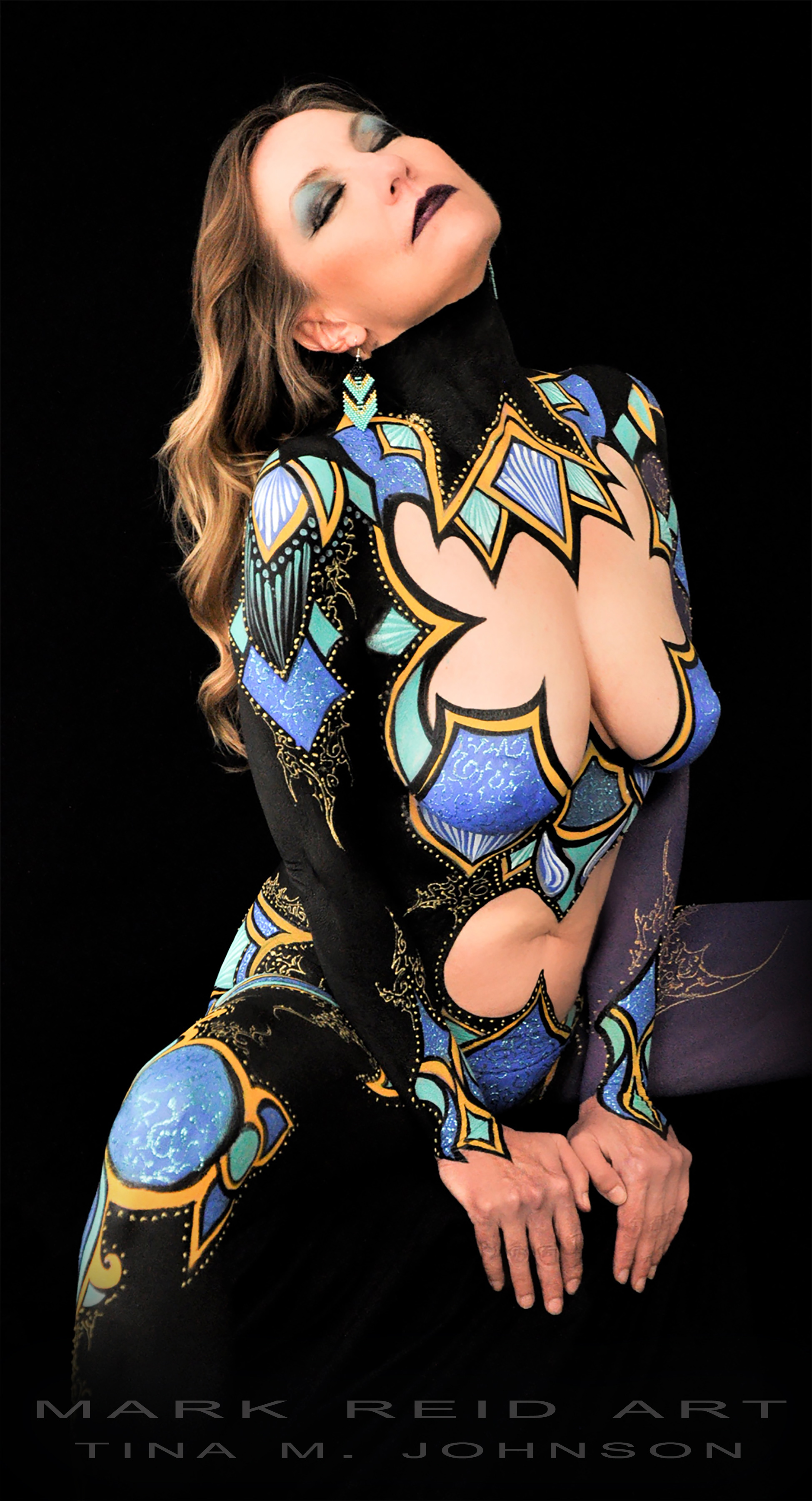 Frontal view of a honey blonde haired woman in full body paint that is brightly colored with combinations of aqua, blue, black and gold paint.  Her eyes are closed and her hands on the front of the stool with her head tilted back.